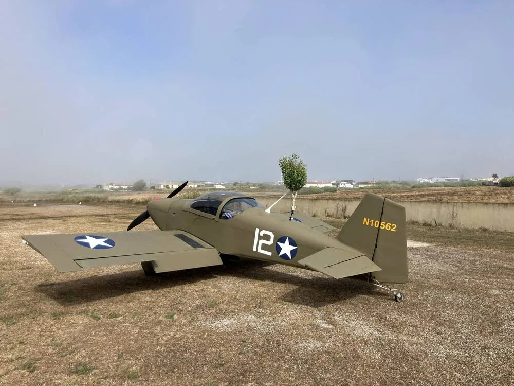 A green single-engine aircraft with a red nose and "N10562" on the tail, parked on a dry grassy field under a hazy sky.