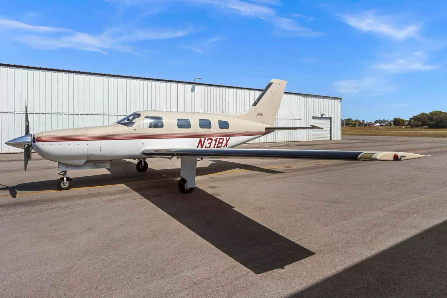 A Piper Malibu aircraft, N31BX, in white and tan with red stripes, parked on an airfield next to a hangar.