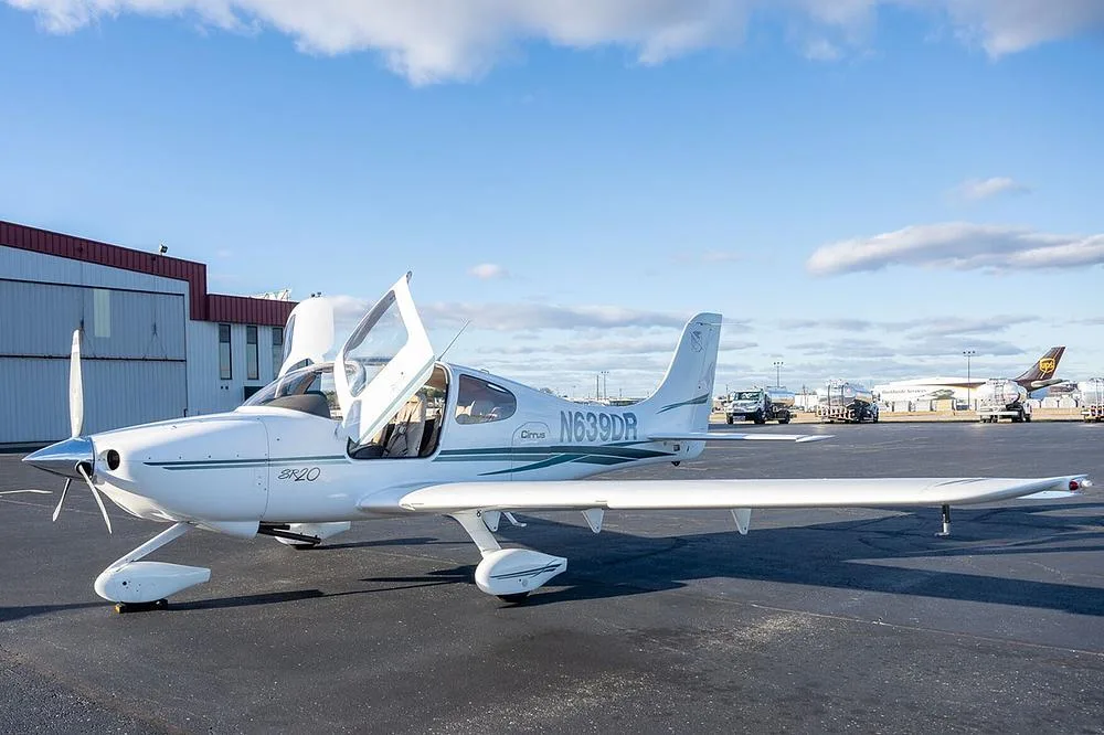 A white Cirrus SR20 aircraft with green and grey stripes, registration N639DR, parked on an asphalt tarmac with its gull-wing doors open.