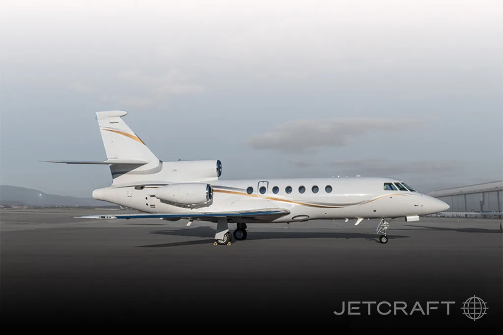 A white Dassault Falcon 900 business jet with gold and grey stripes parked on an airfield.
