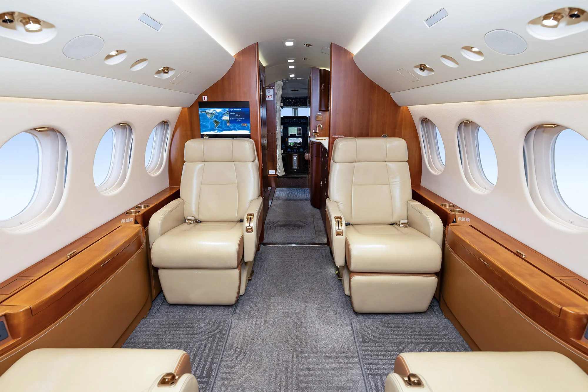 The elegant cabin interior of a business jet, showing beige leather seats, wood paneling, and a monitor displaying a world map.