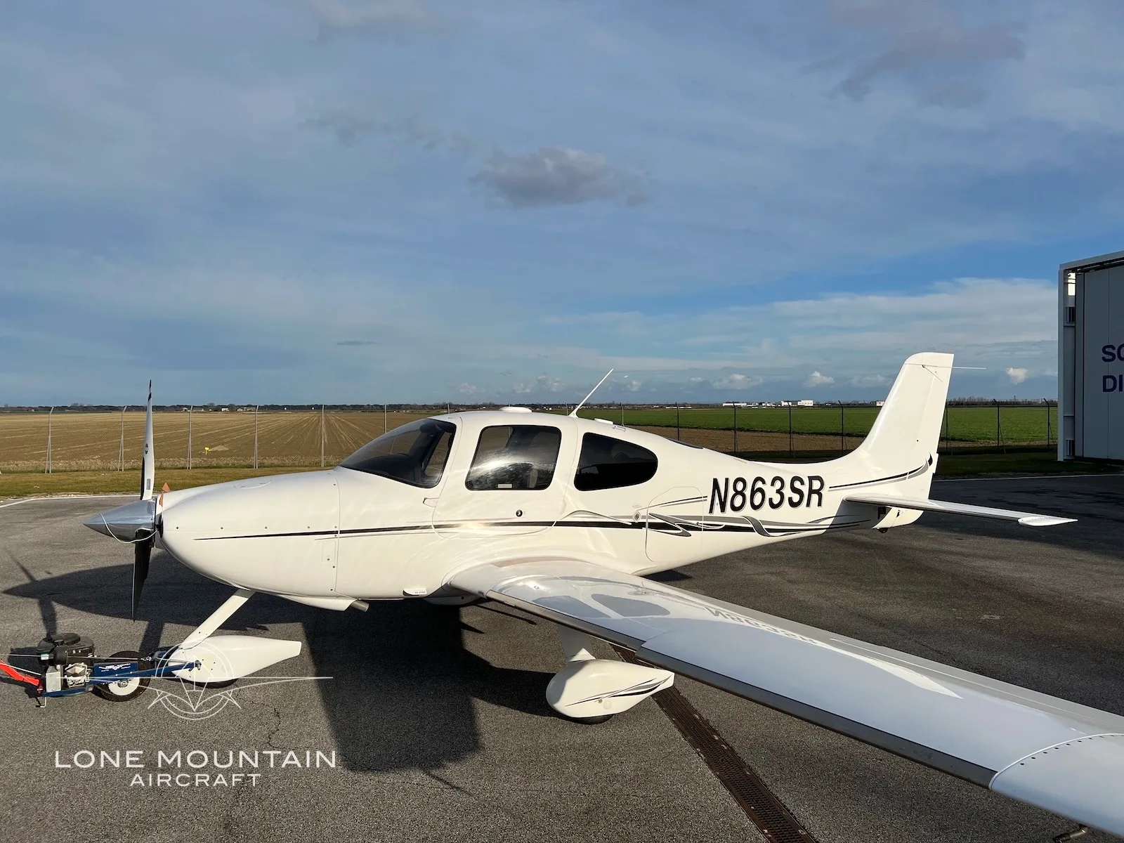 A white Cirrus SR22 aircraft with registration N863SR is parked on an airfield under a partly cloudy sky, with a hangar in the background.