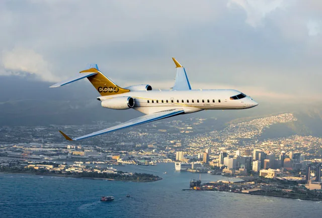A white and gold Bombardier Global business jet flies over a coastal city with mountains in the background.