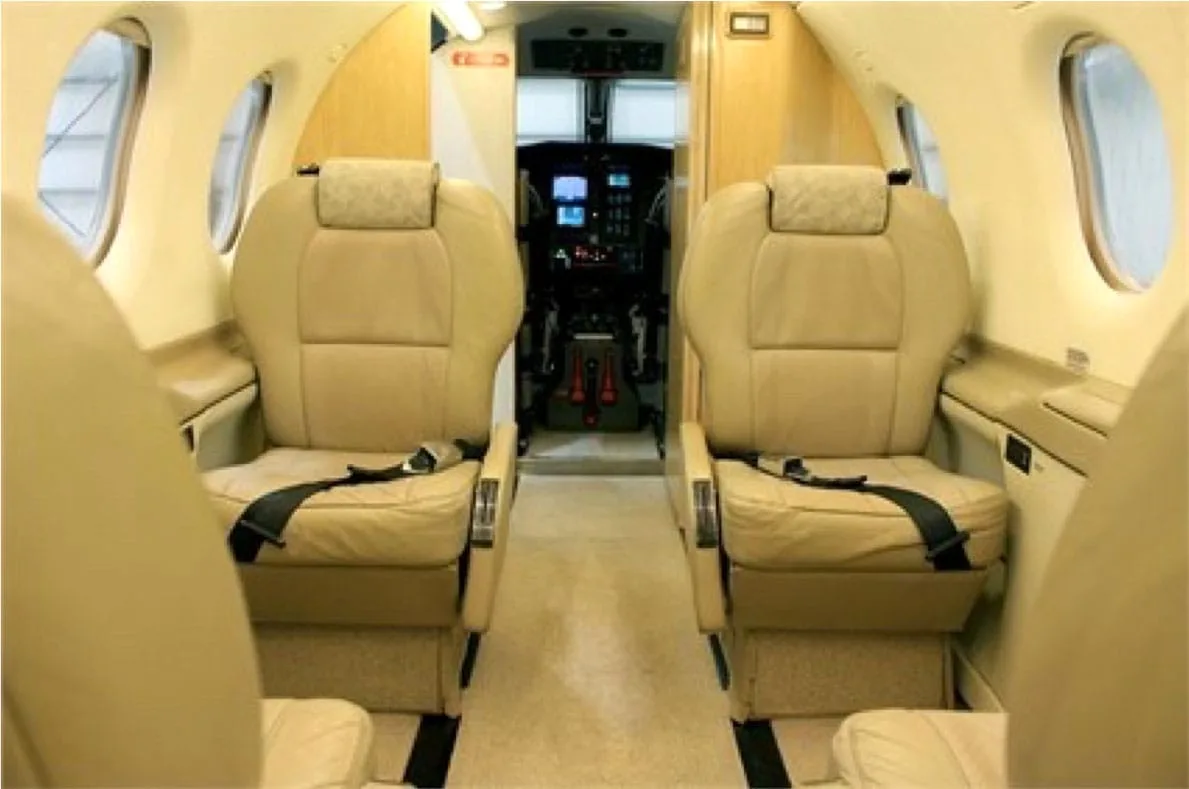 The aircraft cabin interior features two rows of beige leather seats facing forward, with a view towards the cockpit.