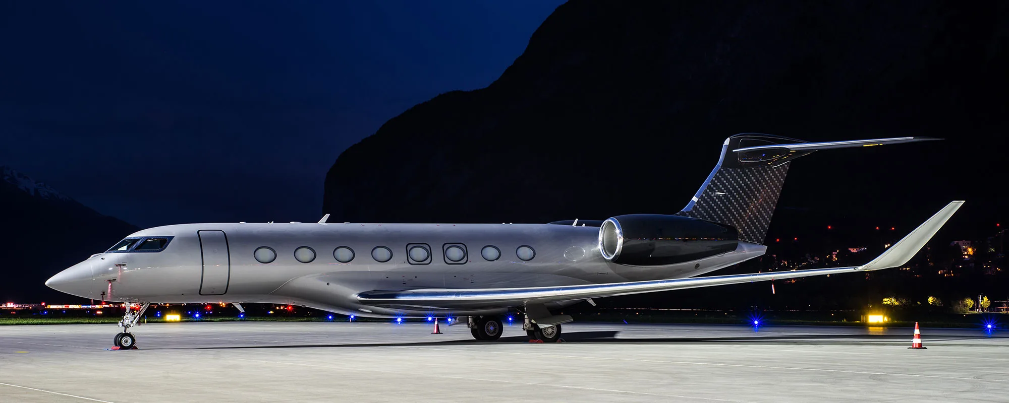A sleek silver and black business jet is parked on an airport tarmac at night, with mountains in the background.