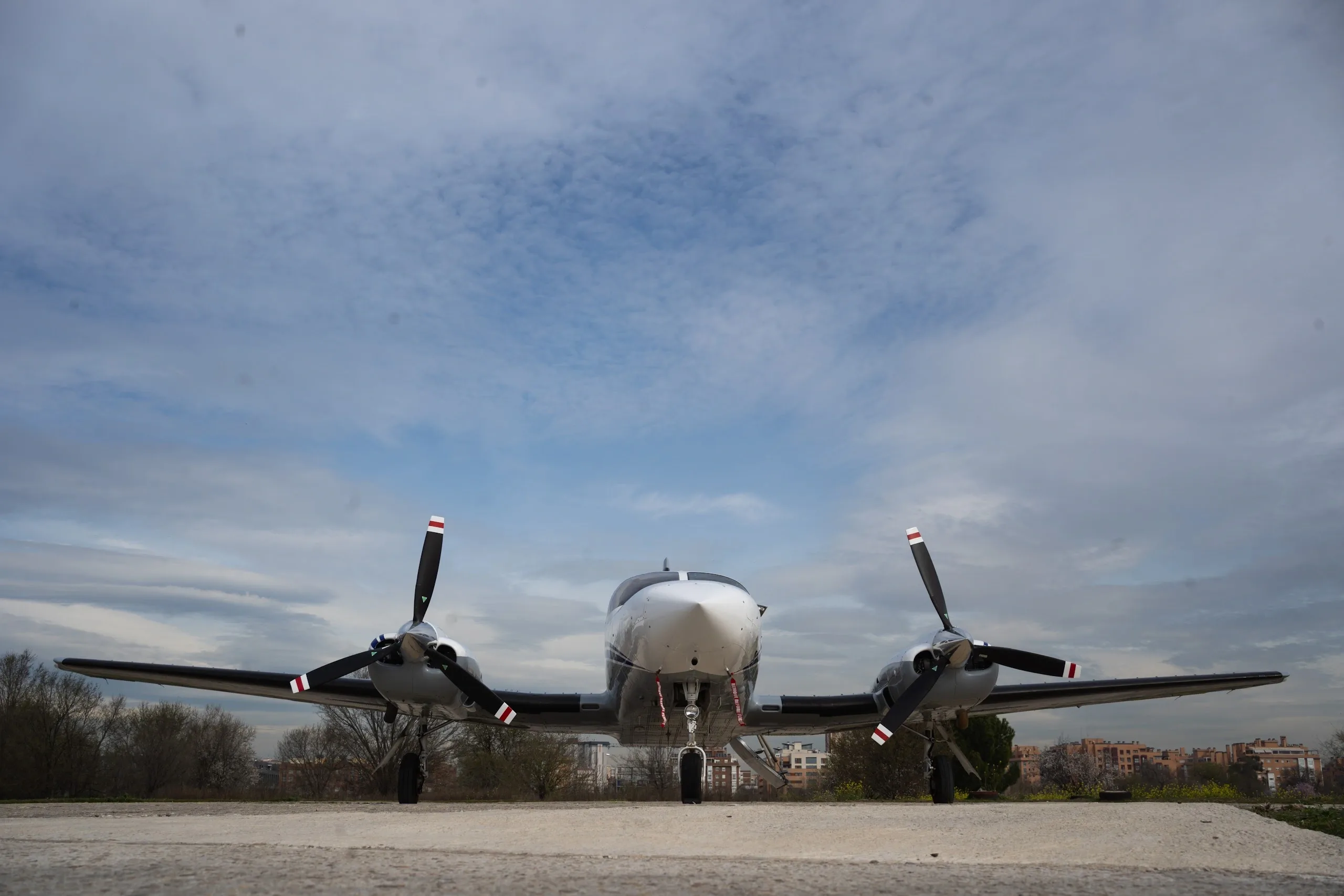 A low-angle front view of a white and grey twin-engine propeller aircraft with blue stripes on a concrete pad under a partly cloudy sky.
