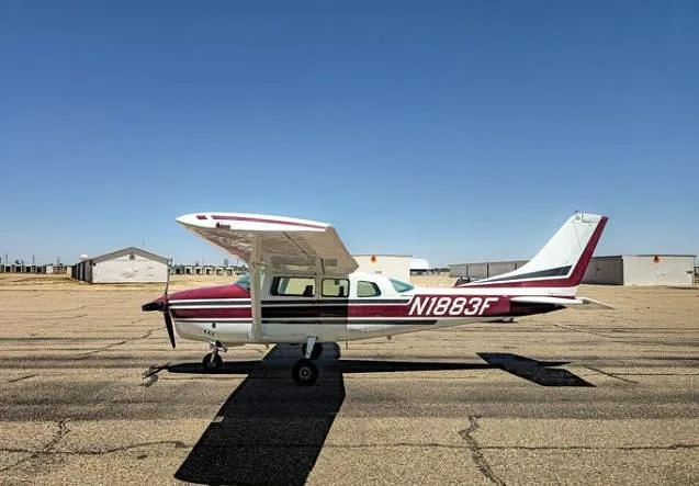 A side view of a white and maroon Cessna 210 Centurion aircraft with registration N1883F, showing its high wing design, parked on an airfield.