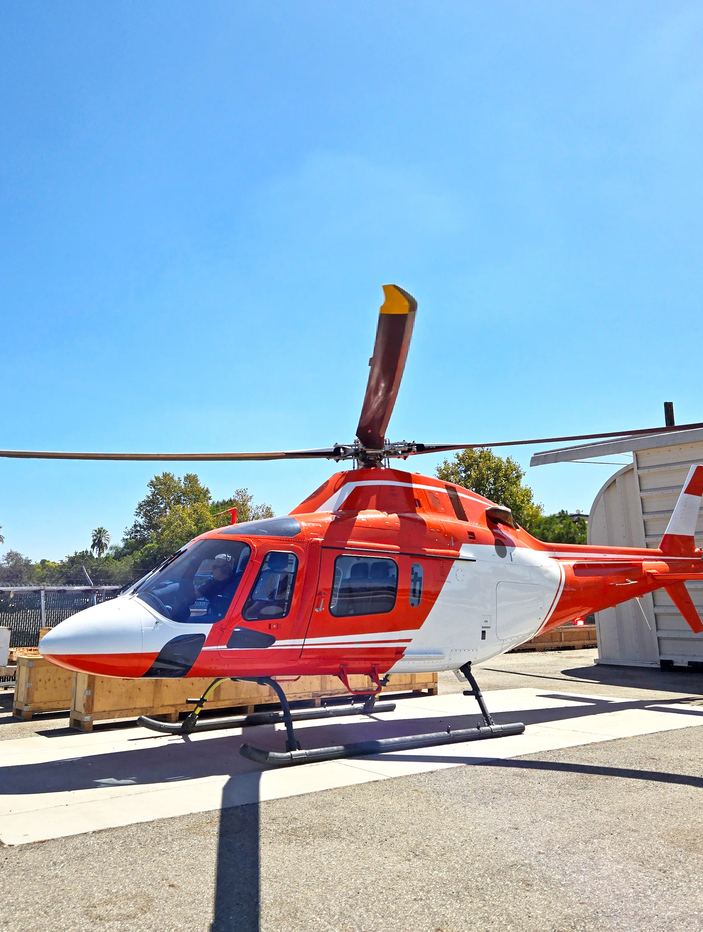 A red and white helicopter with a white nose and tail, parked on an asphalt surface under a clear blue sky.