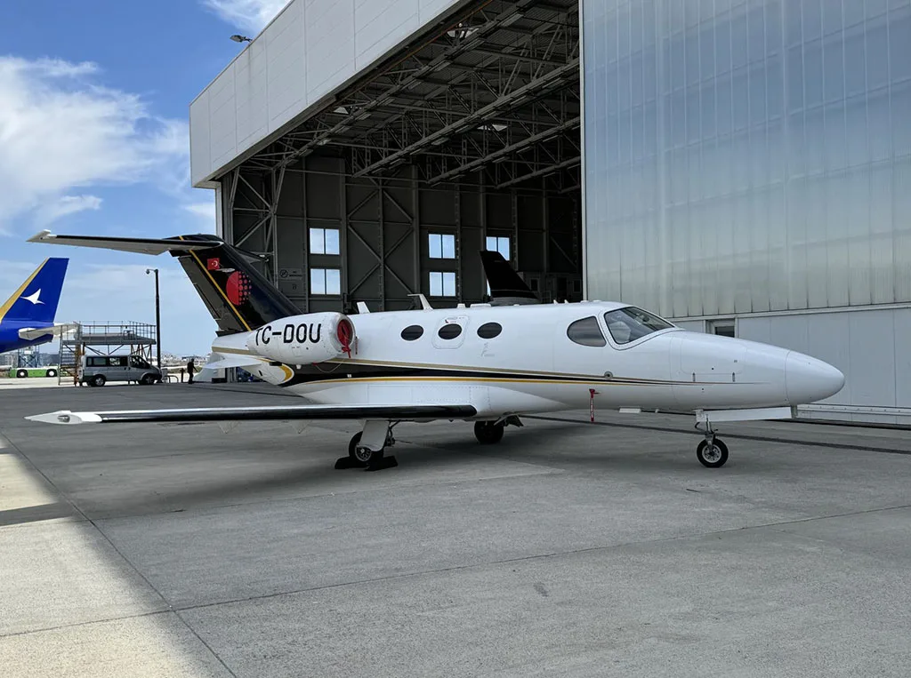 A white business jet with black and gold accents on the tail, registration TC-OOU, parked on an airport apron next to a large hangar.