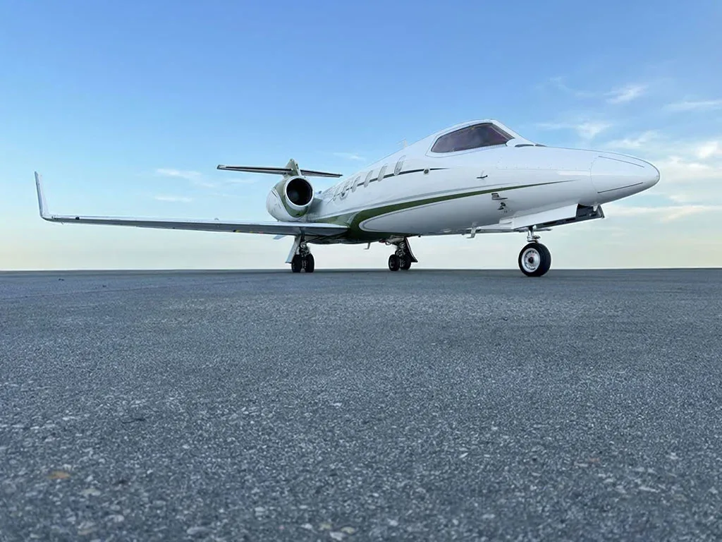 A white and green Learjet 31A (N784AM) is parked on an asphalt runway under a blue sky.