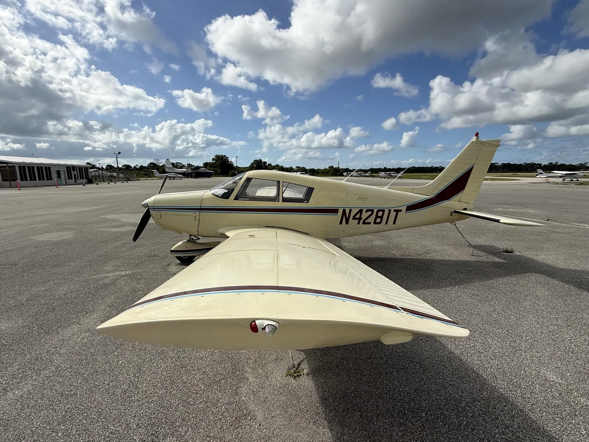 A cream-colored Piper PA-28-140E Cherokee with maroon and blue stripes, registration N4281T, parked on an airfield under a partly cloudy sky.