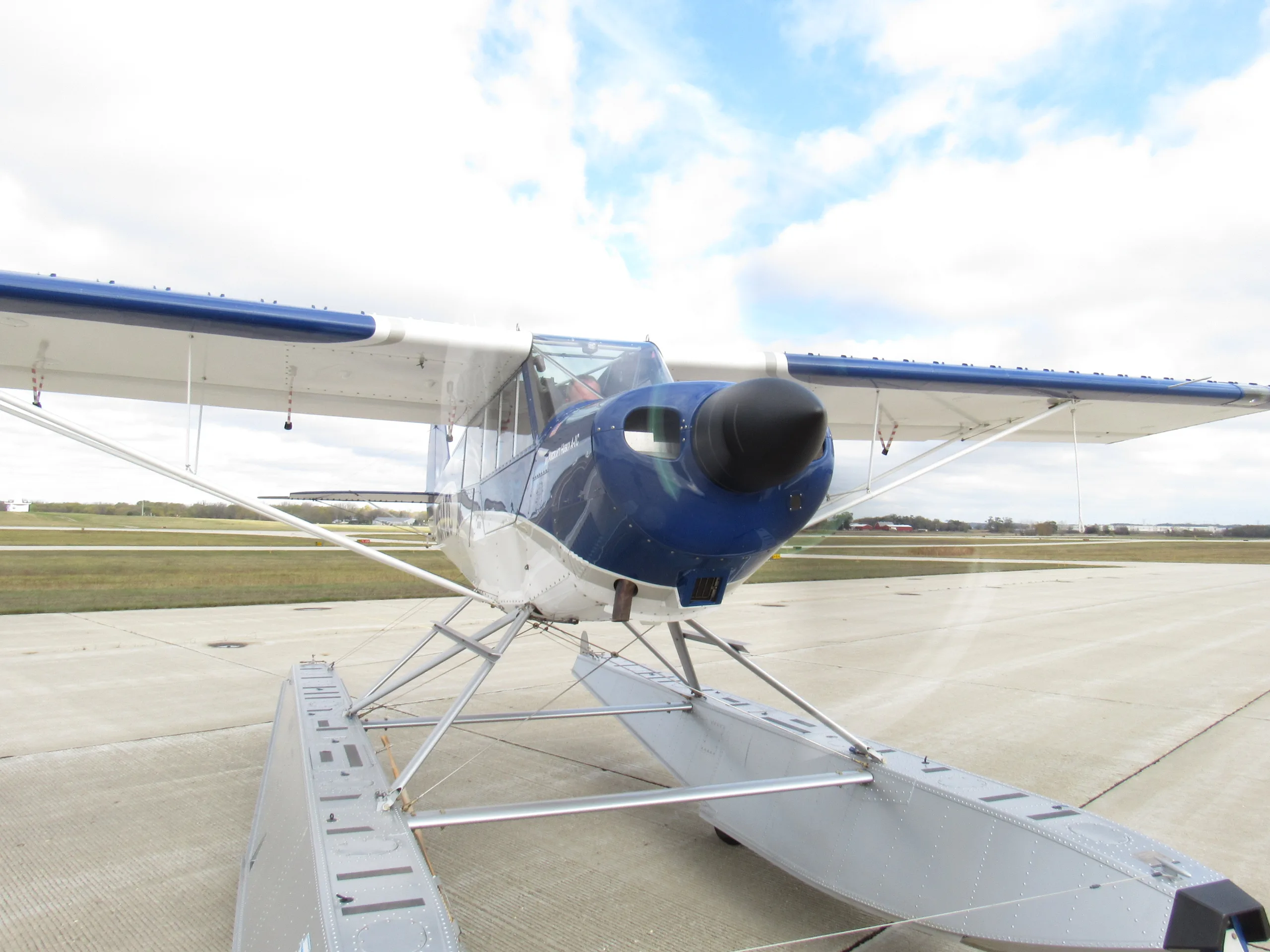A blue and white floatplane with large silver floats is parked on a concrete airfield under a cloudy sky, viewed from a low angle.