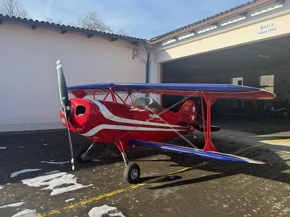 A red Pitts Special biplane with blue upper wings and white stripes on the lower wings, parked in front of a hangar.