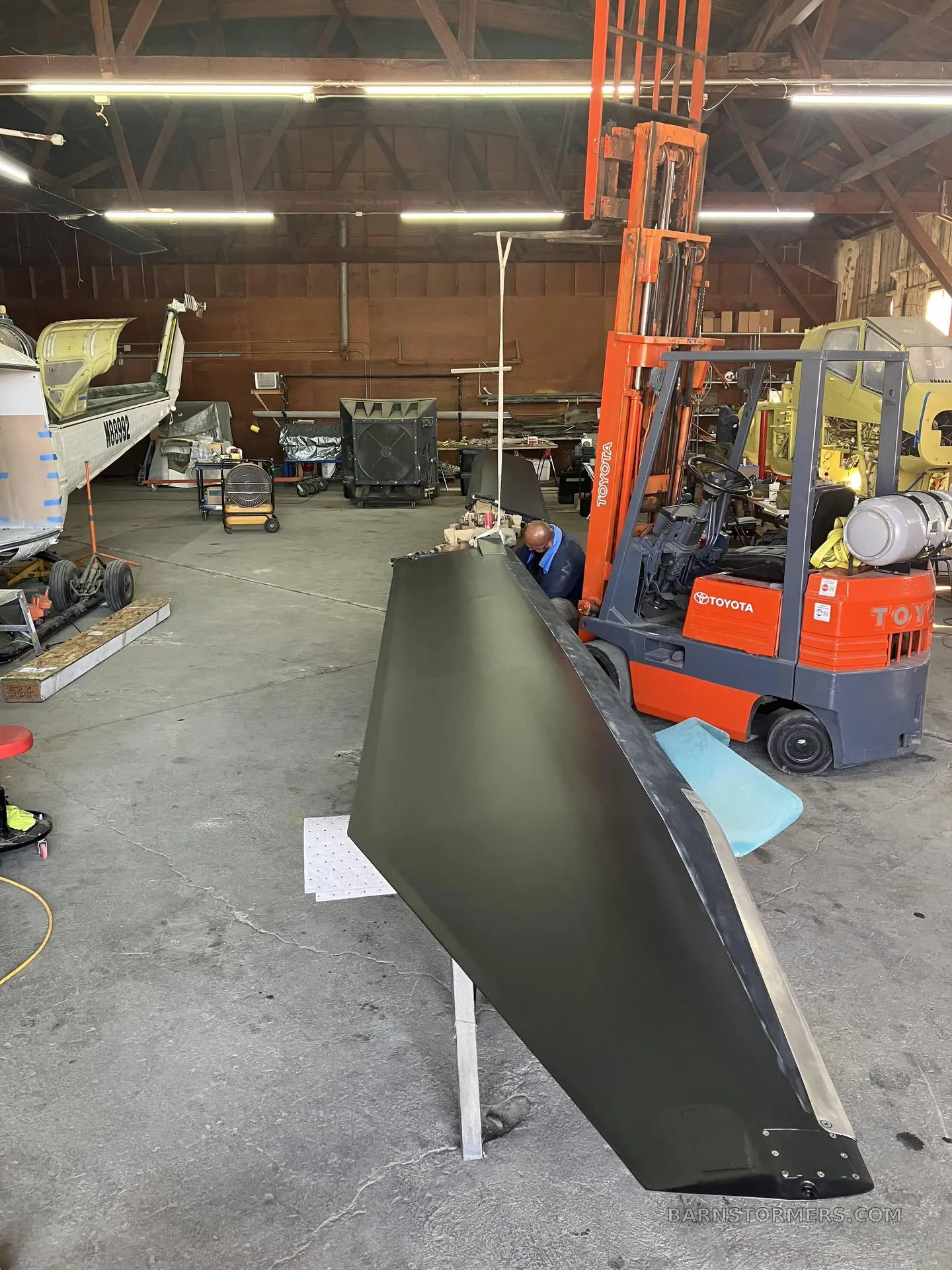 A dark green aircraft component, possibly a tail section, is being worked on in a hangar with a forklift nearby.
