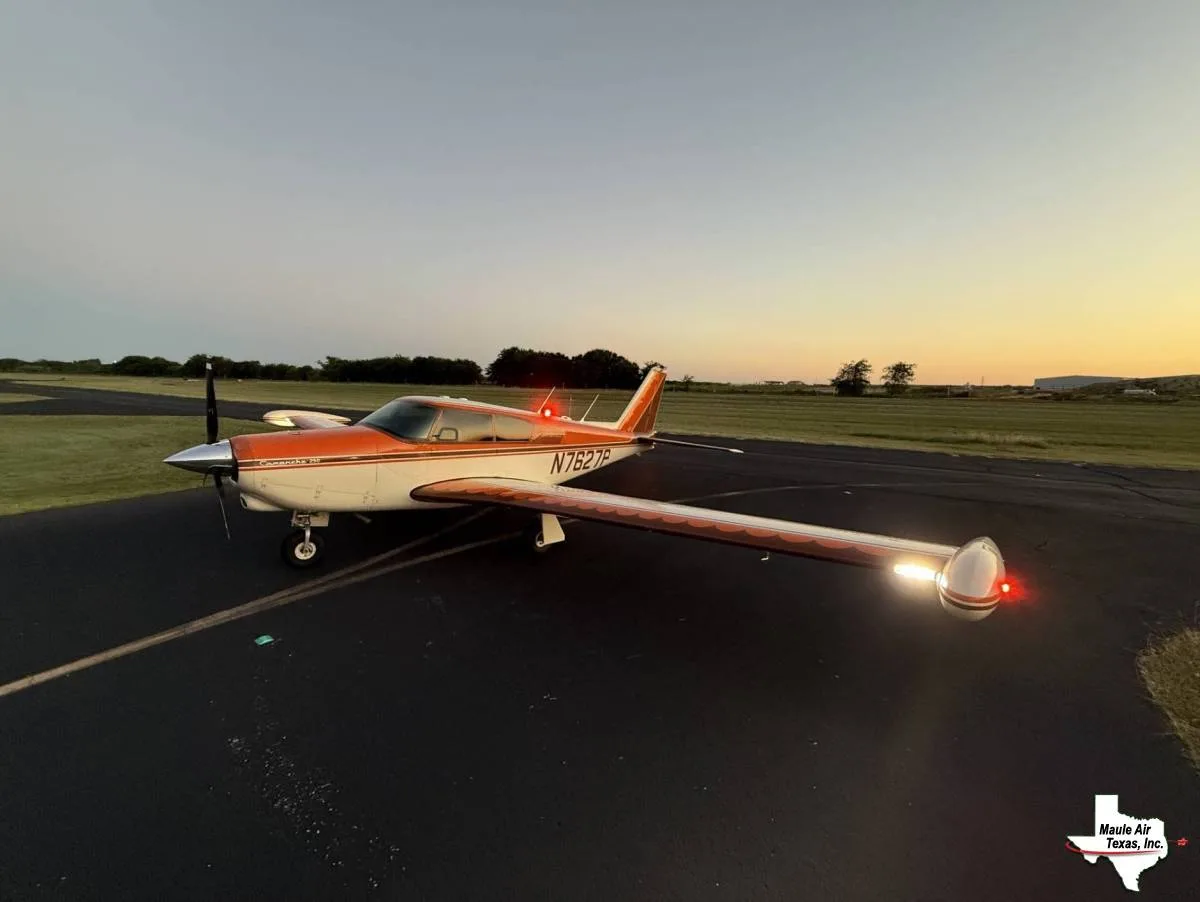 A Piper Comanche 250 aircraft, painted orange and white, with navigation lights illuminated, parked on an airfield at dusk.