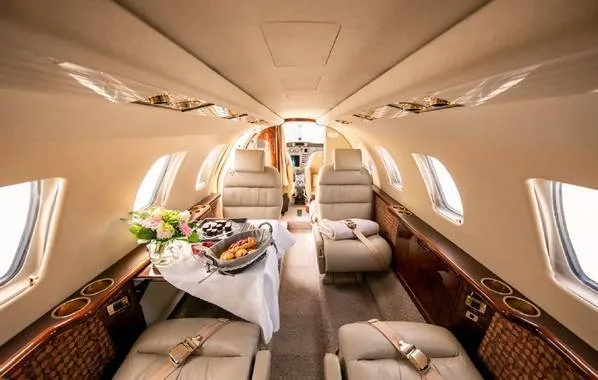 The luxurious interior cabin of a private jet, featuring beige leather seats and a table set with food and flowers.
