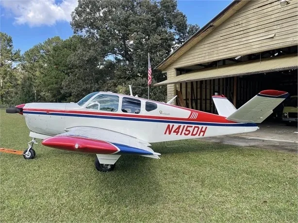 A side view of a white and red Beechcraft Bonanza N415DH parked on grass next to a wooden hangar.