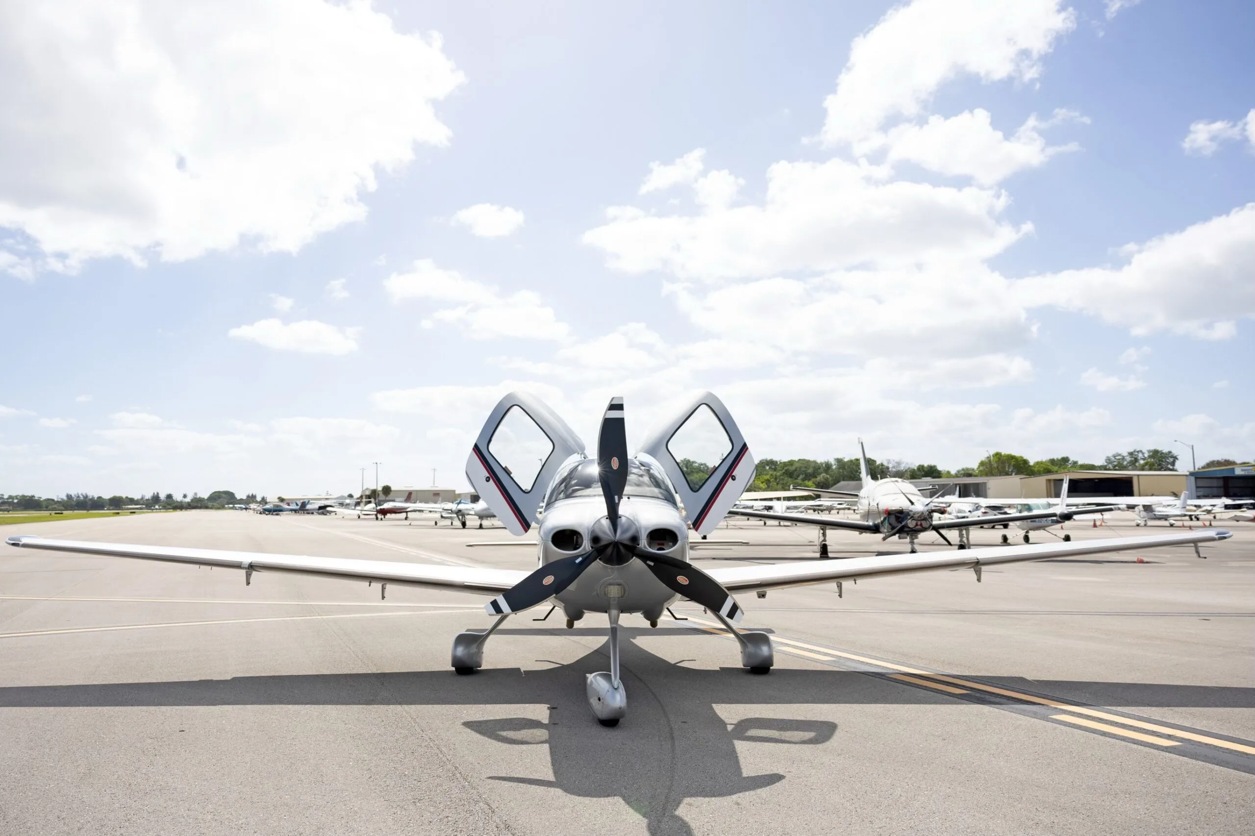 A head-on view of a silver Cirrus SR22 GTS aircraft with doors open, parked on an airfield under a partly cloudy sky.
