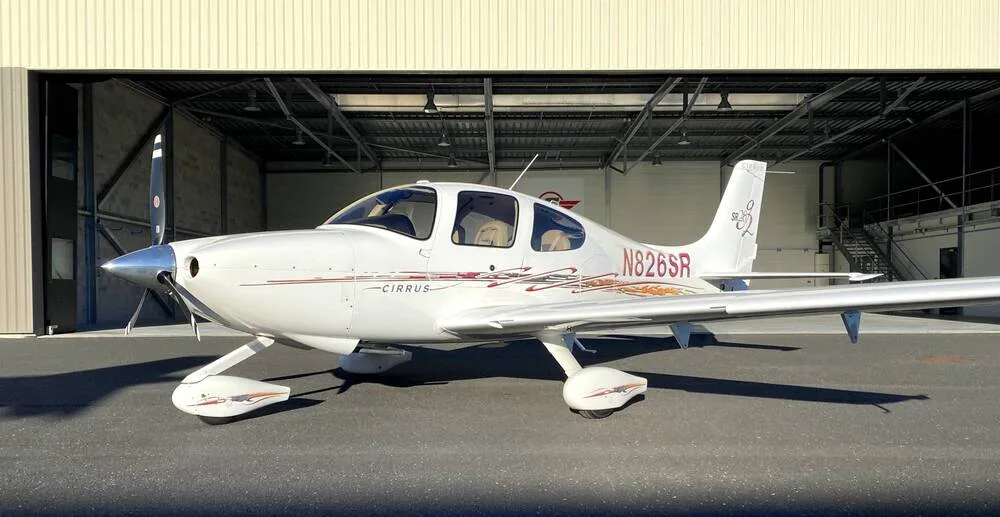 A three-quarter front view of a white Cirrus SR22 aircraft with red and gold accent stripes, registration N826SR, parked in front of an open hangar.