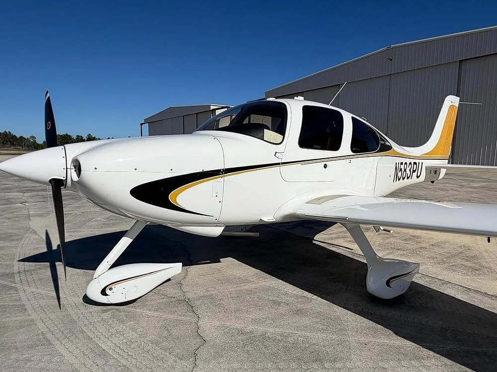 A close-up front-left view of a white Cirrus SR22 aircraft with black and gold stripes, registration N583PU, parked near hangars.