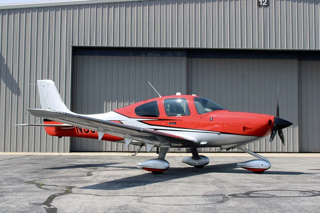 A red and silver Cirrus SR22 aircraft with registration N394JM parked on an tarmac in front of a hangar.