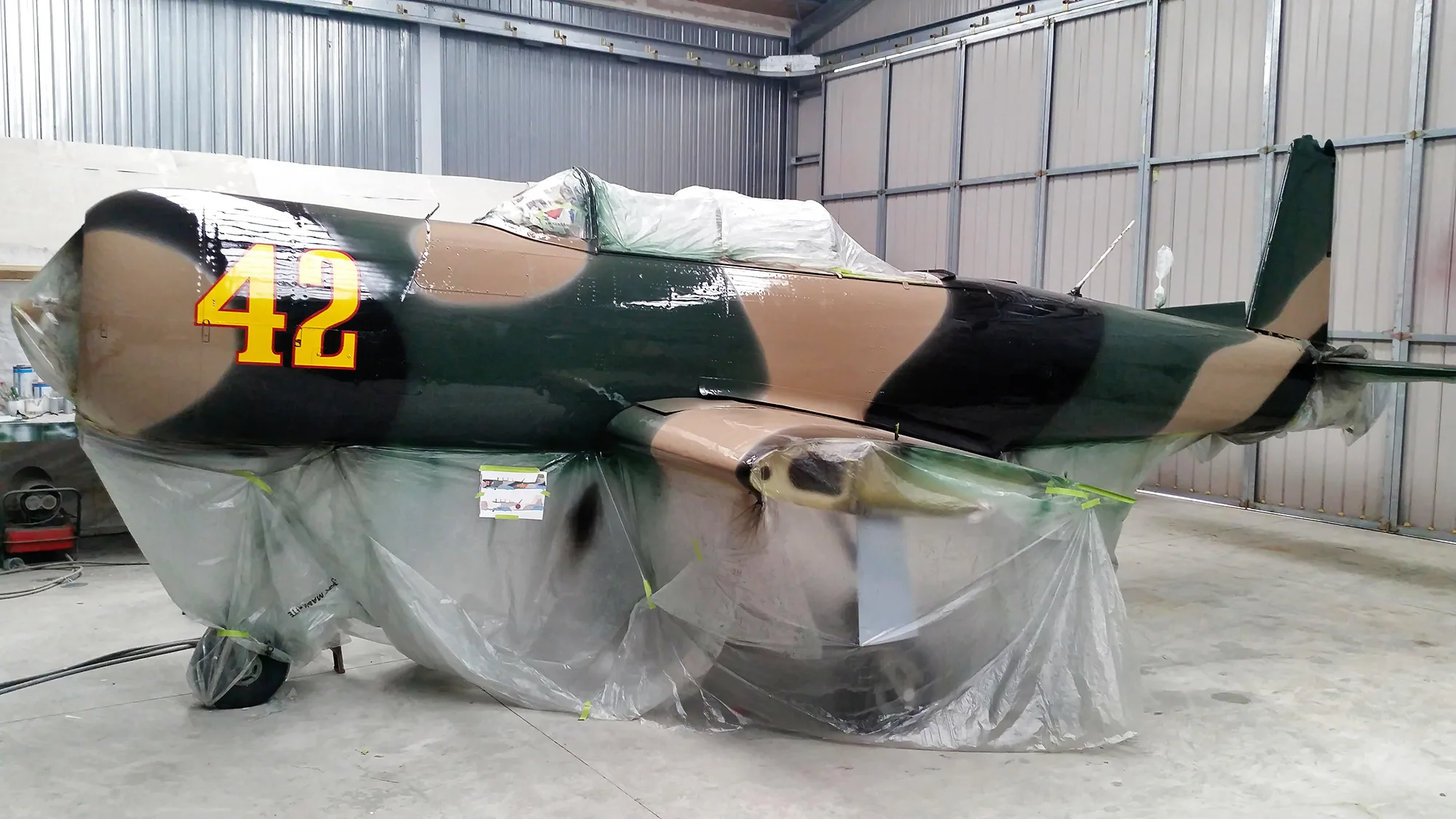 A single-engine aircraft with a camouflage paint scheme (green, brown, black) and the number "42" in yellow on the fuselage, partially covered in plastic sheeting in a hangar.