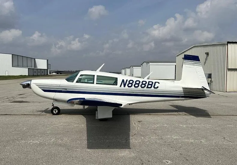 A white single-engine aircraft with blue and grey stripes, registration N888BC, parked on an asphalt ramp with hangars in the background.