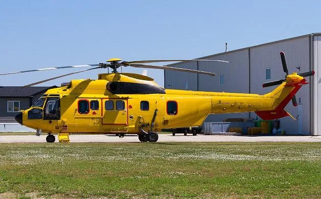A large yellow Airbus H225 Super Puma helicopter with a red tail, parked on an airfield next to a hangar.