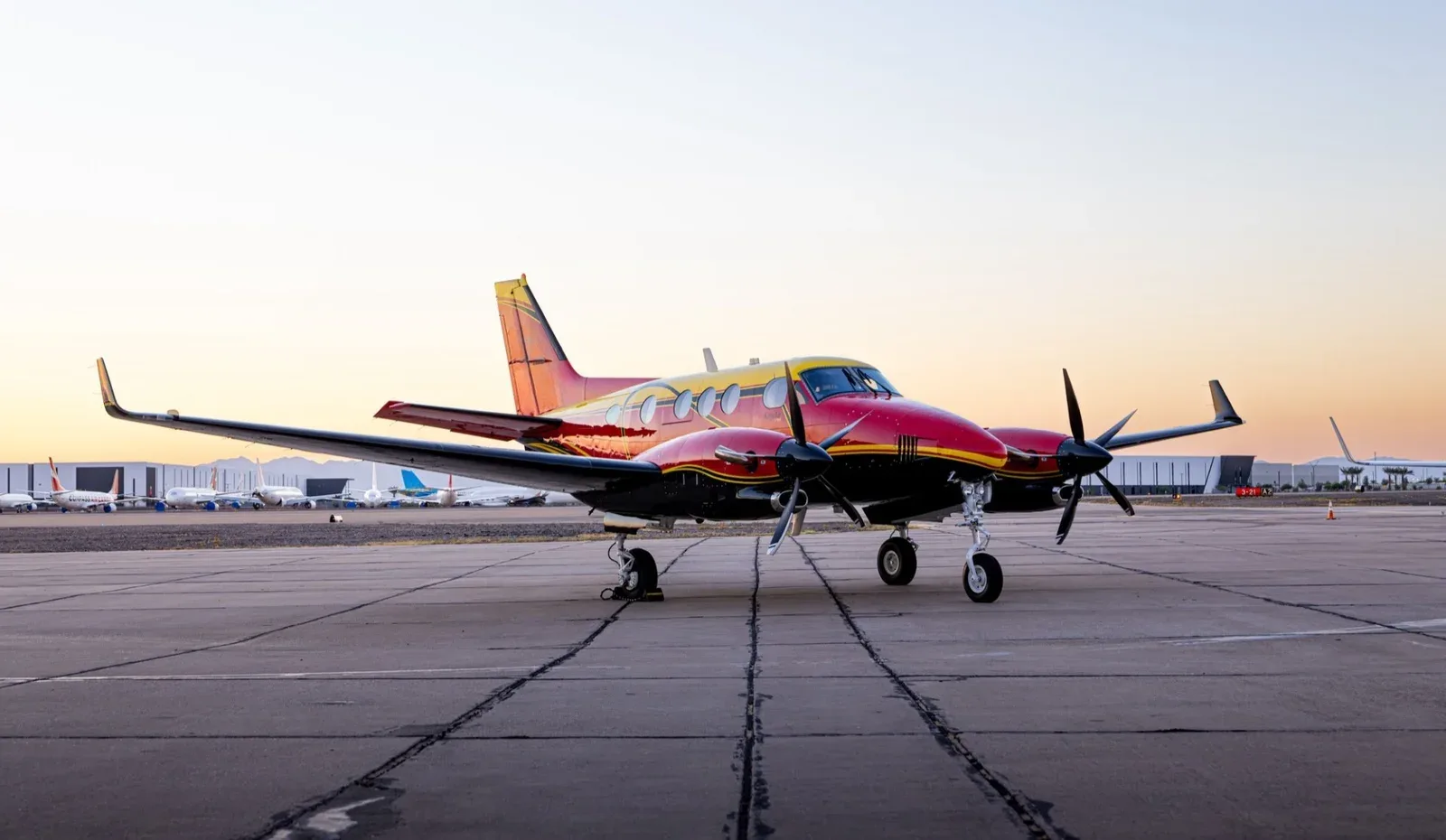 A red, yellow, and black King Air C90B turboprop aircraft parked on an airfield under a clear sky.