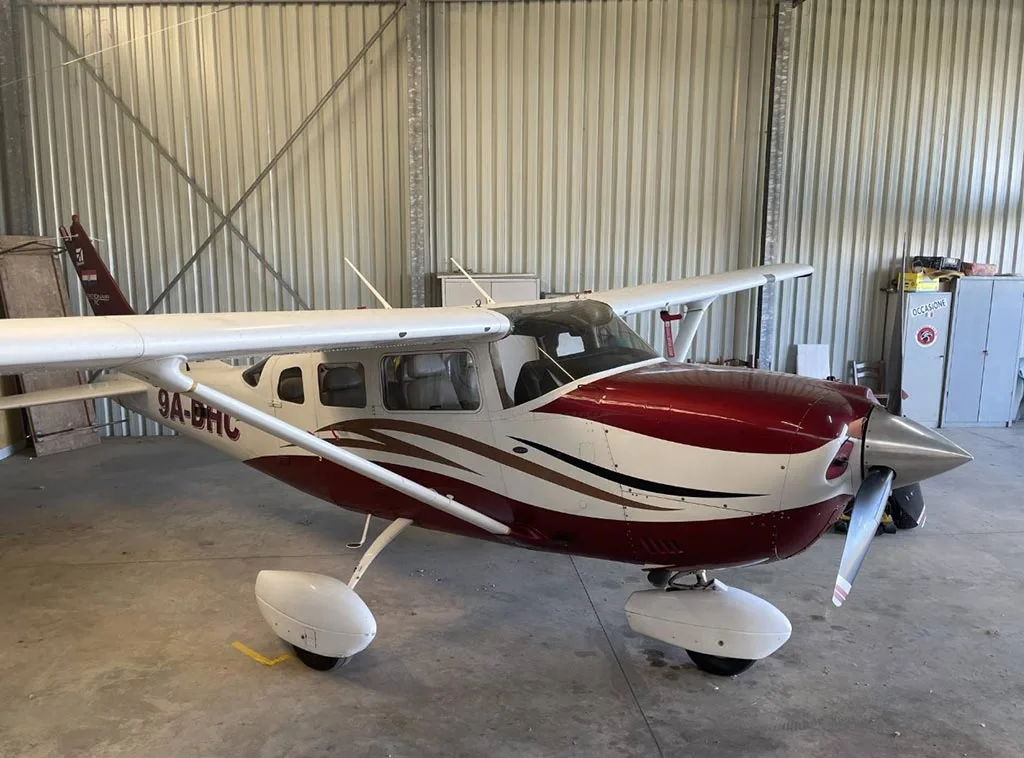 A three-quarter front view of the red and white Cessna 206/210 aircraft 9A-DHC in a hangar.