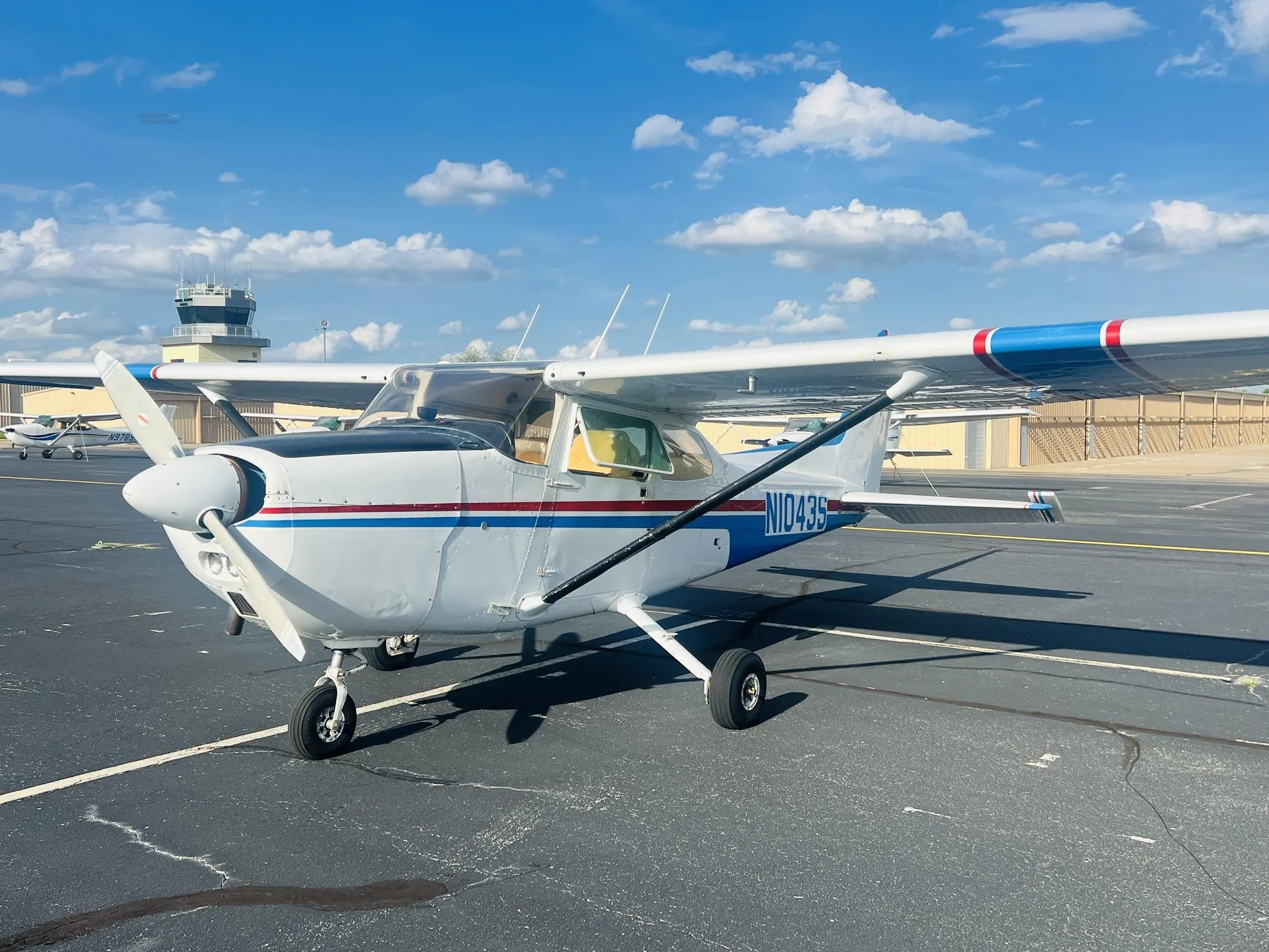 A white single-engine propeller aircraft with blue and red stripes is parked on an asphalt tarmac under a blue sky, with a control tower in the background.