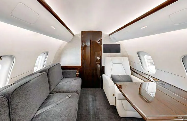 A section of the private jet interior with a grey sofa, a cream armchair, and a large wooden table.