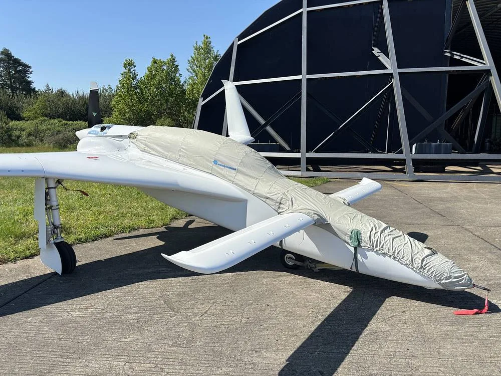 A white, single-engine aircraft, partially covered with a grey cover, is parked on a concrete pad on a sunny day.