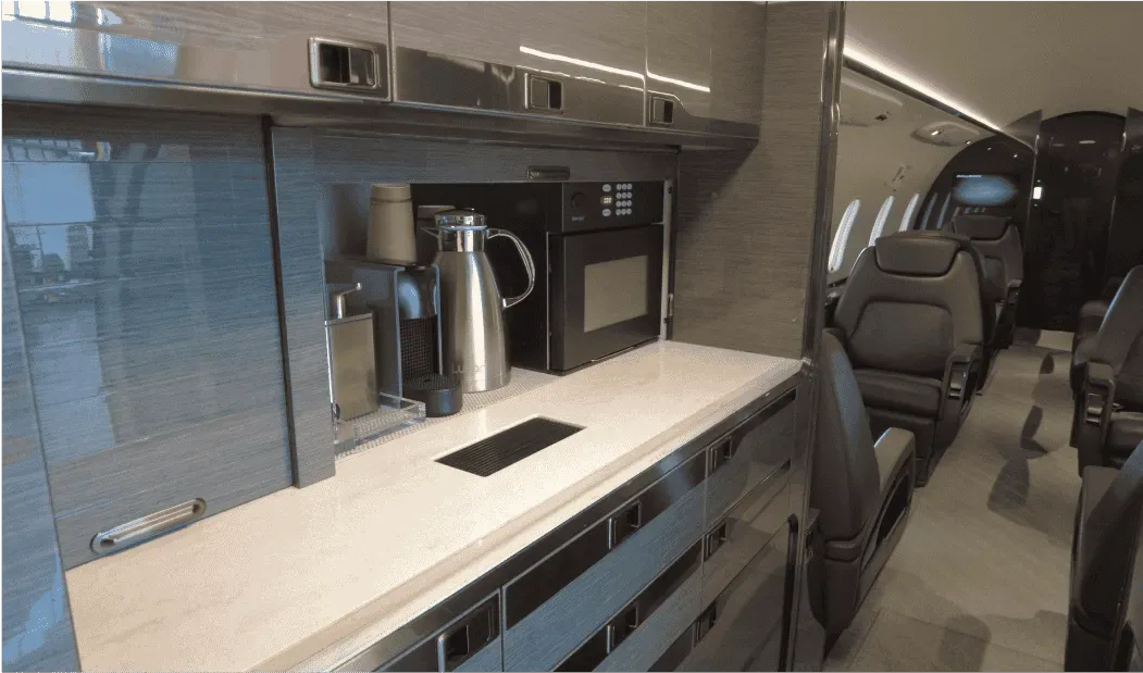 The galley area of a business jet cabin featuring a countertop, coffee maker, and microwave, with passenger seats visible in the background.