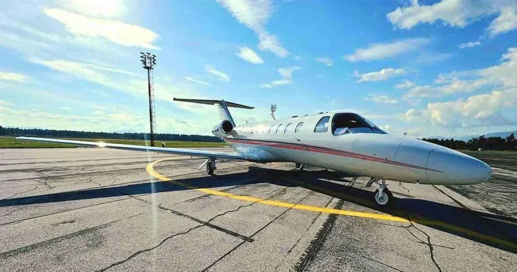 A white Cessna CitationJet with red stripes parked on an asphalt runway, viewed from a lower angle with sun glare.