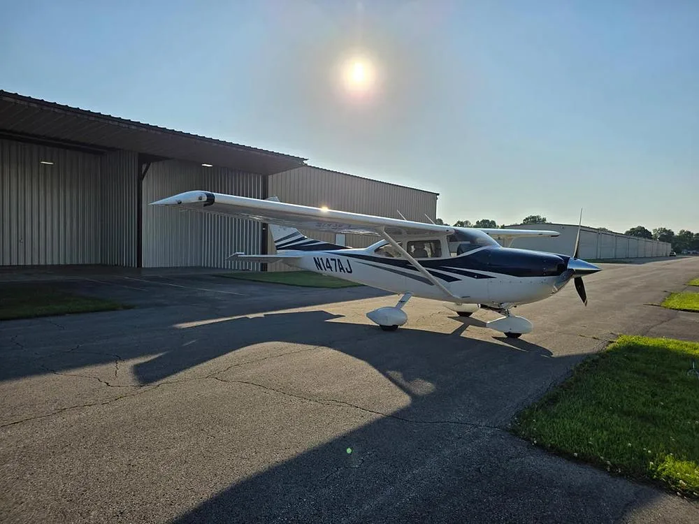 A white and blue Cessna 182 aircraft with registration N147AJ parked on an asphalt ramp with hangars in the background and sun glare.