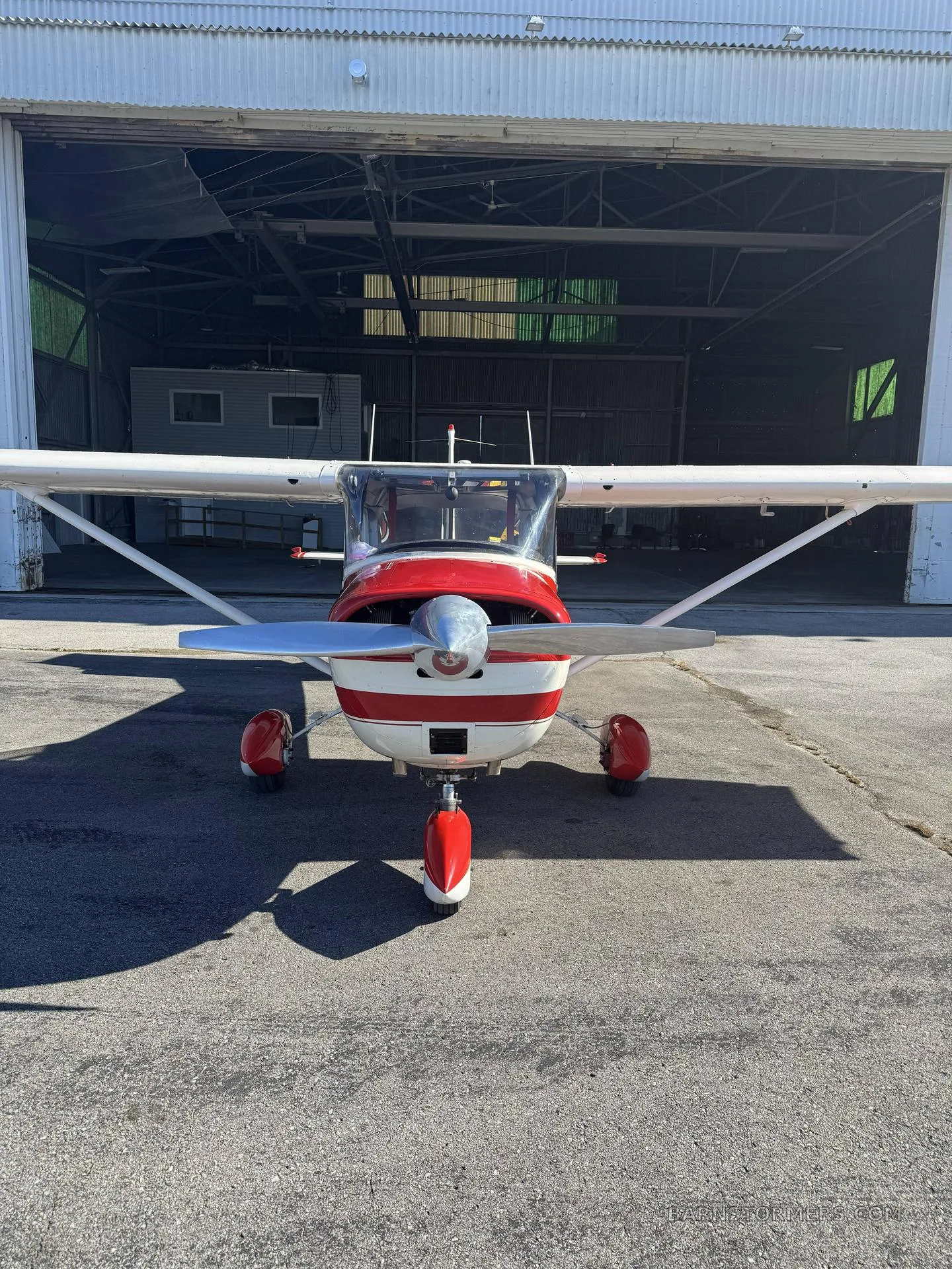A red and white single-engine aircraft with a propeller, viewed head-on outside a hangar.