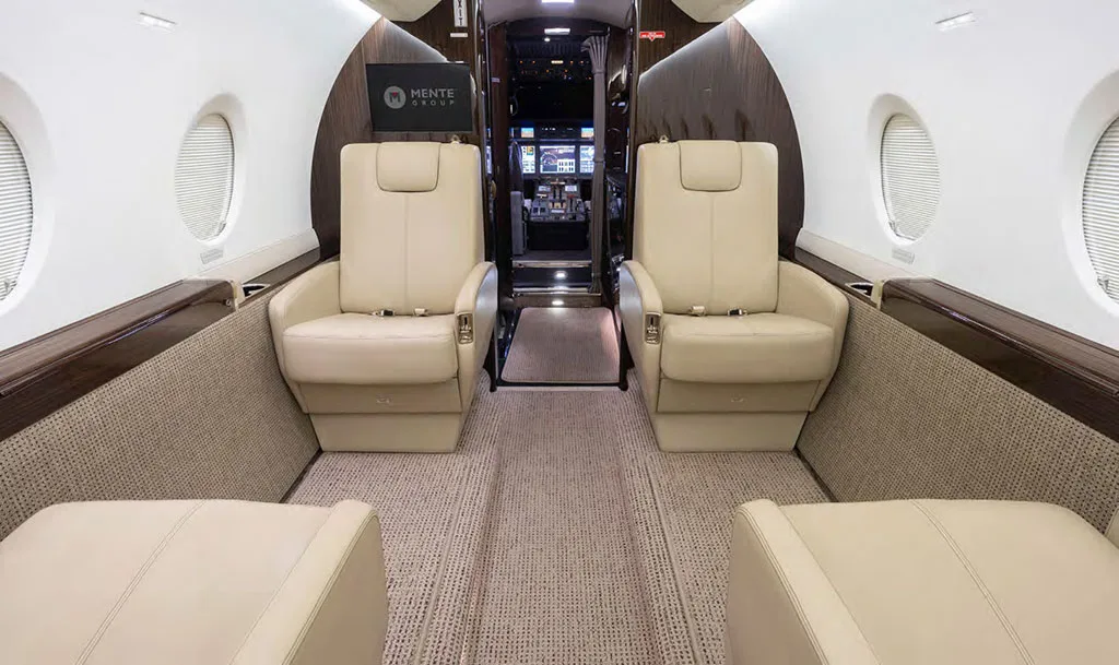 The luxurious interior cabin of the Gulfstream G550 features light-colored leather seats and dark wood accents, looking towards the cockpit.