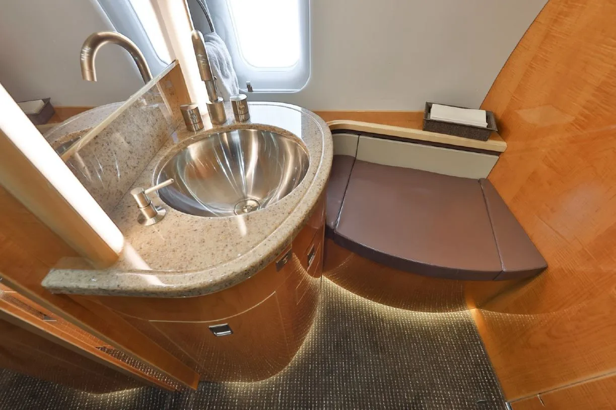 The aircraft lavatory features a modern sink with a polished metal basin and elegant wood and granite-like finishes.