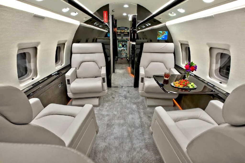 The luxurious interior cabin of a private jet, featuring light gray leather seats, a dark table with fruit, and a galley in the background.