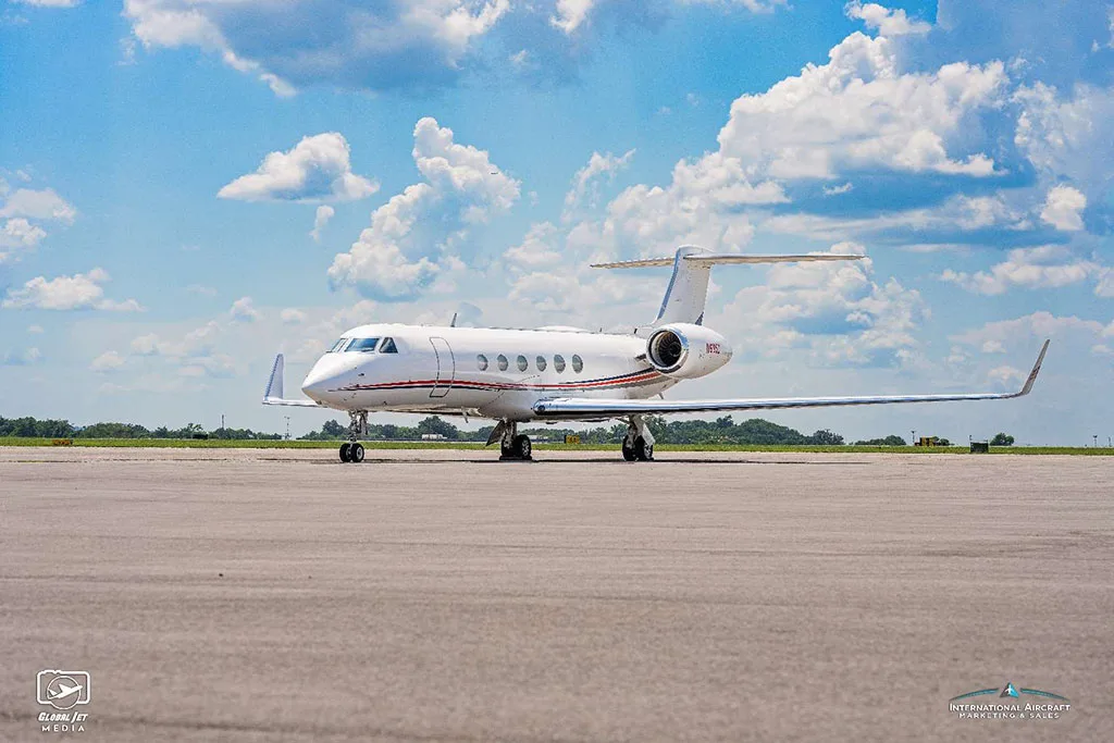 A side view of a white Gulfstream G550 aircraft with red and blue stripes, parked on an airfield under a blue sky.