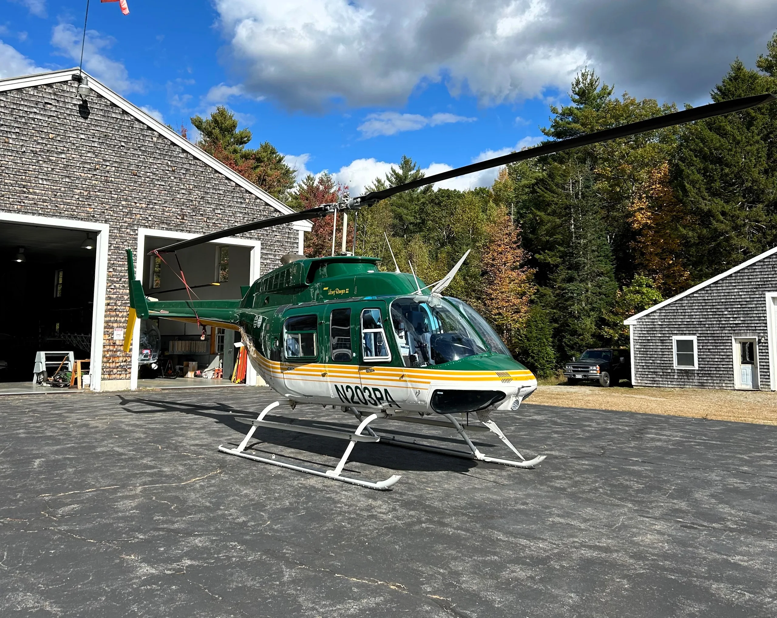 A green and white Bell 206L-3 LongRanger III helicopter, registration N203PA, parked on an asphalt pad outside a shingled hangar on a sunny day.