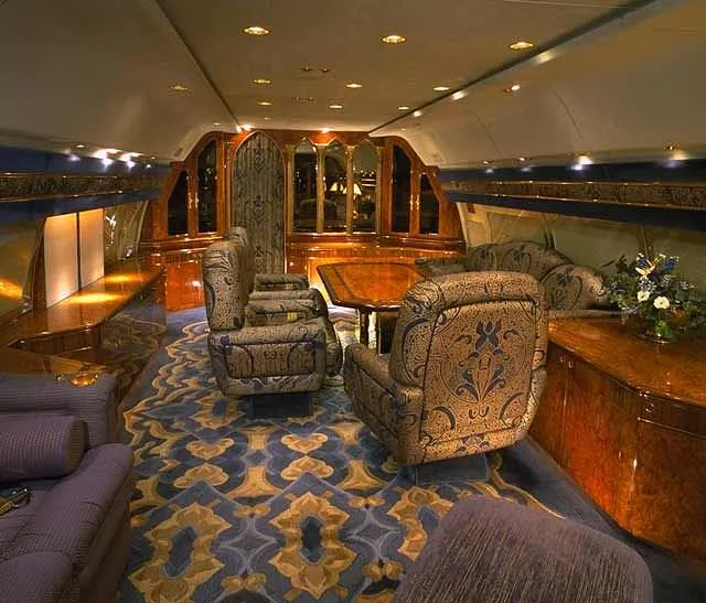 A luxurious aircraft interior features ornate blue and gold patterned carpet, plush seating, and polished wood cabinetry.