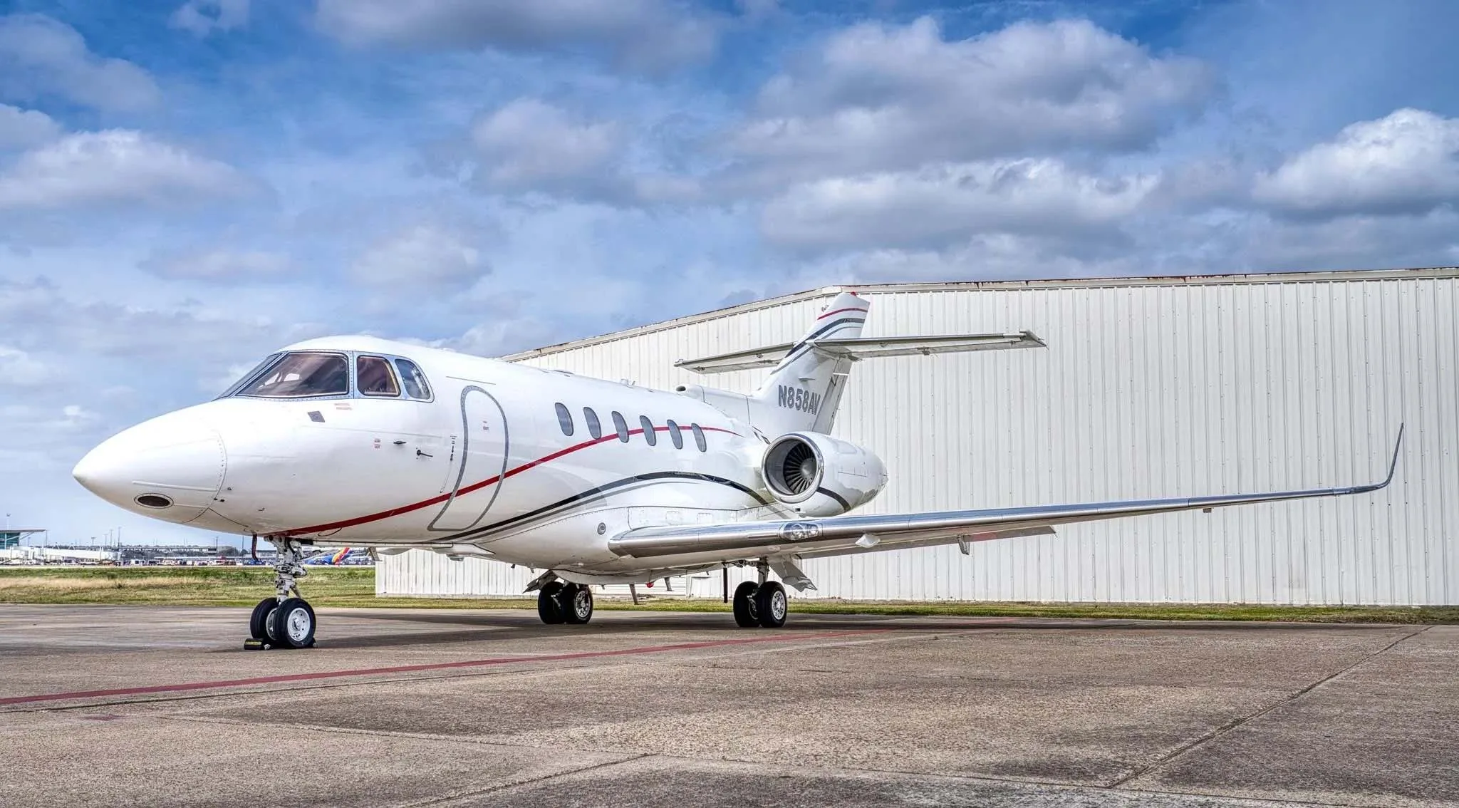 A white Dassault Falcon 900 business jet with red and black stripes parked on an airfield in front of a hangar.