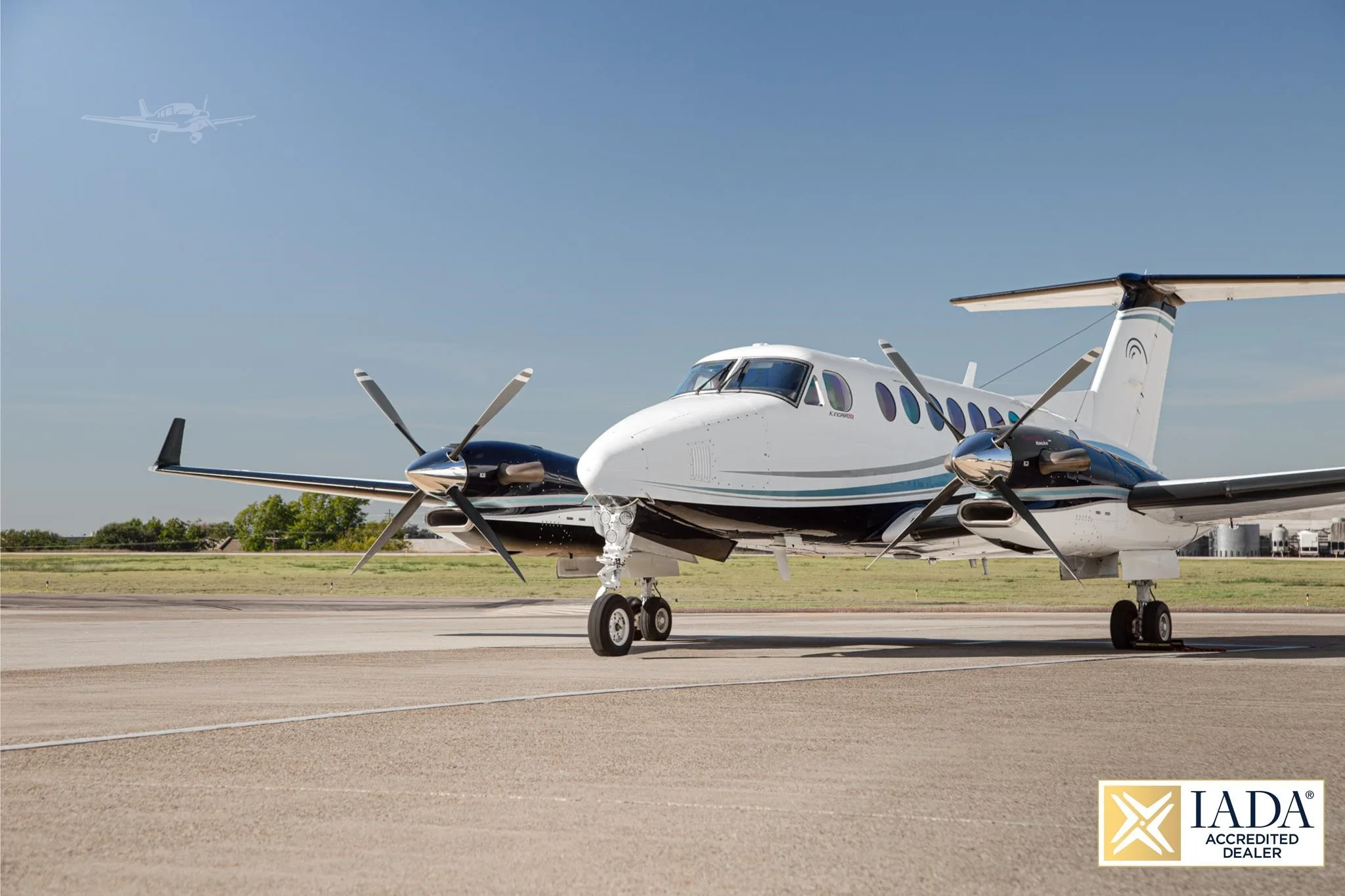 A white and black Beechcraft King Air twin-turboprop with a blue stripe, parked on a tarmac under a clear blue sky.