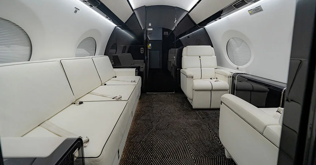 The business jet cabin interior featuring a white leather sofa, individual seats, and dark patterned carpet.