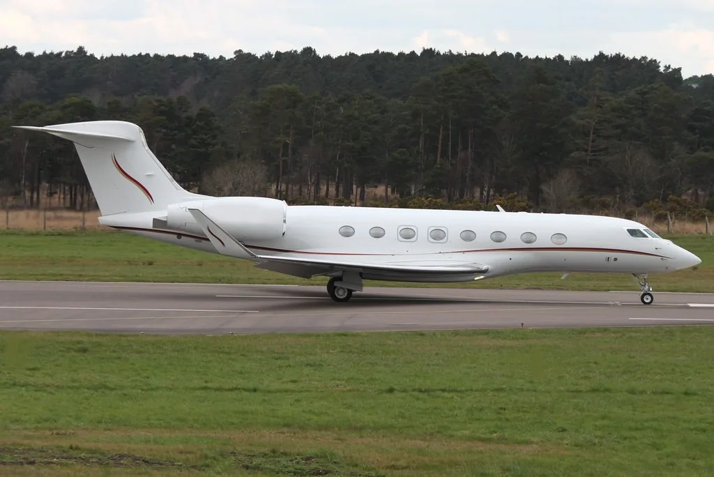 A white Gulfstream business jet with red and gold stripes on the tail and fuselage, on a runway with grass and trees in the background.