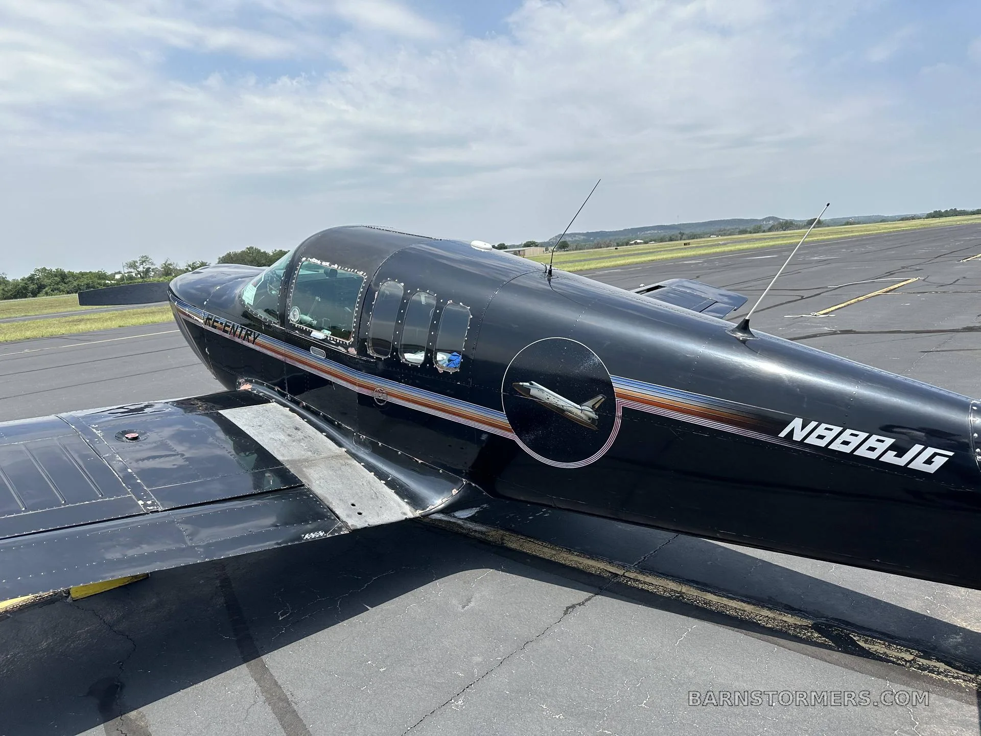 A black single-engine aircraft with 'RE-ENTRY' and registration N888JG on its side, parked on an asphalt runway under a partly cloudy sky.