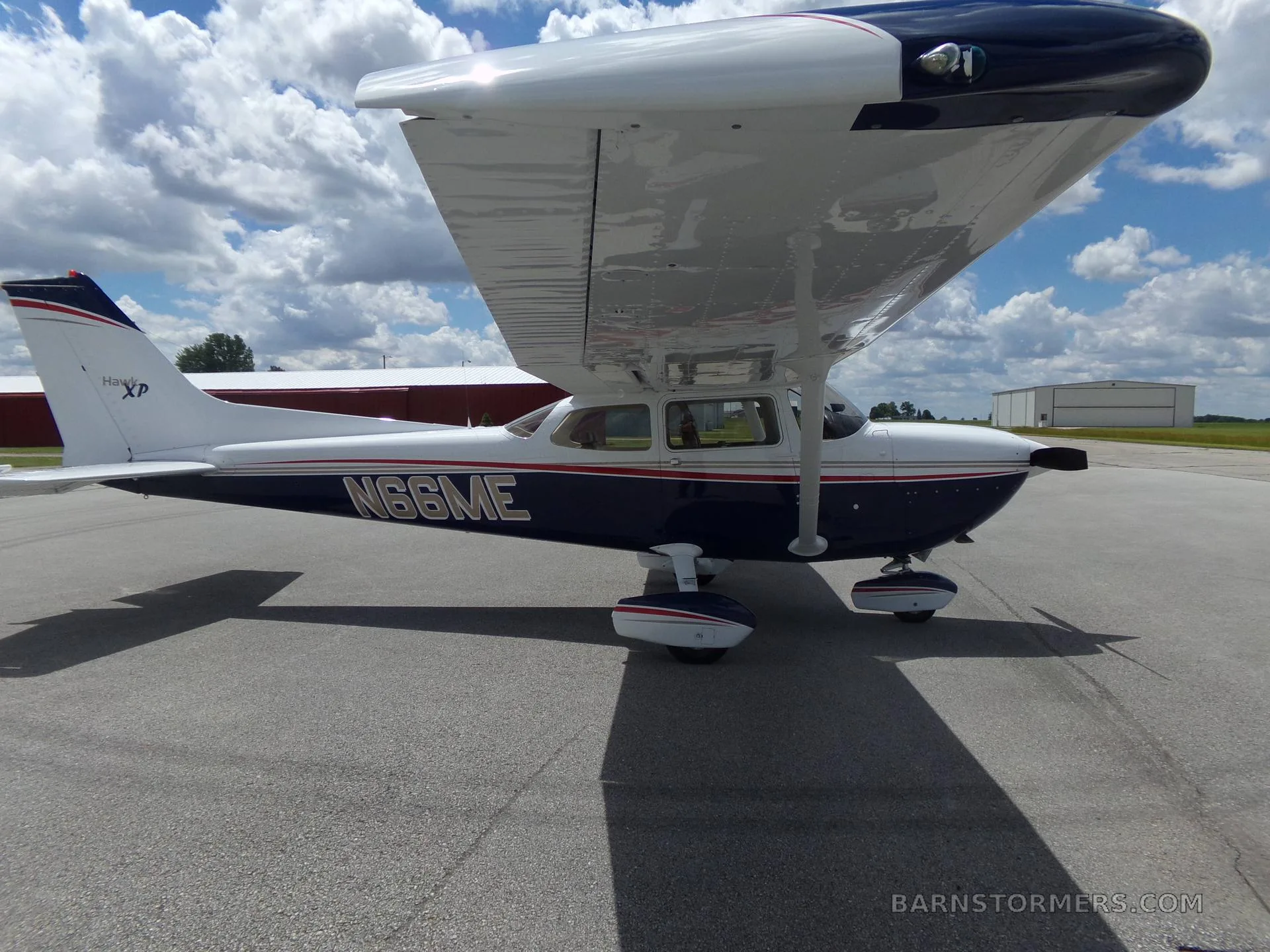 A white and blue Cessna 172 Hawk XP with red and gold stripes, seen from the side-right on an airfield with hangars in the background.