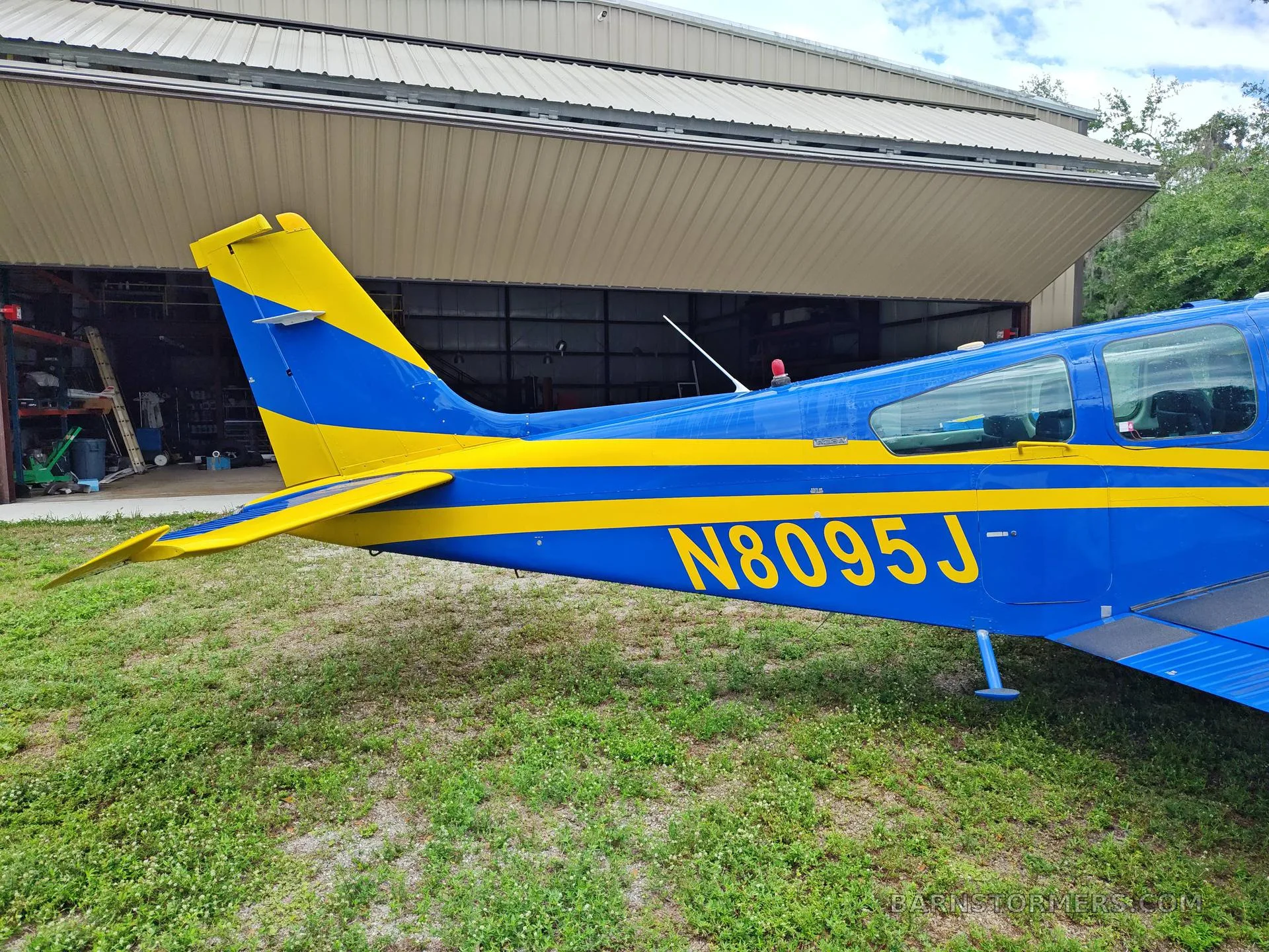 A close-up side view of the blue and yellow tail and rear fuselage of a single-engine aircraft, with a hangar in the background.